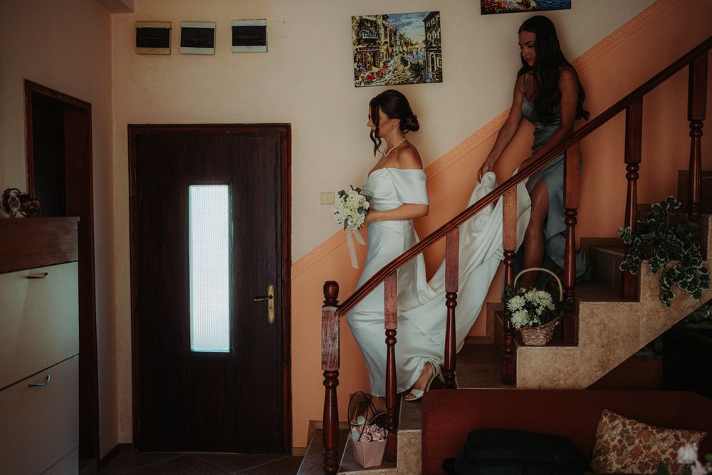 Two people in formal attire descend a wooden staircase in a home interior.