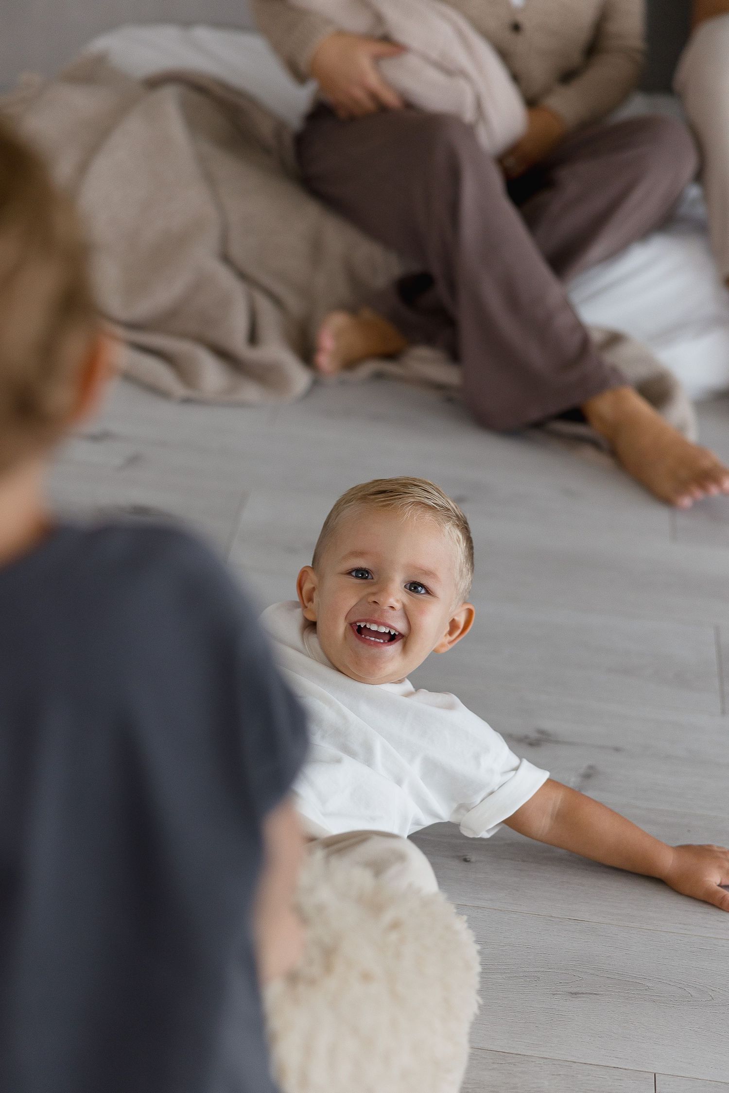 A smiling baby wearing white clothing lies on a light gray floor while playing with others.