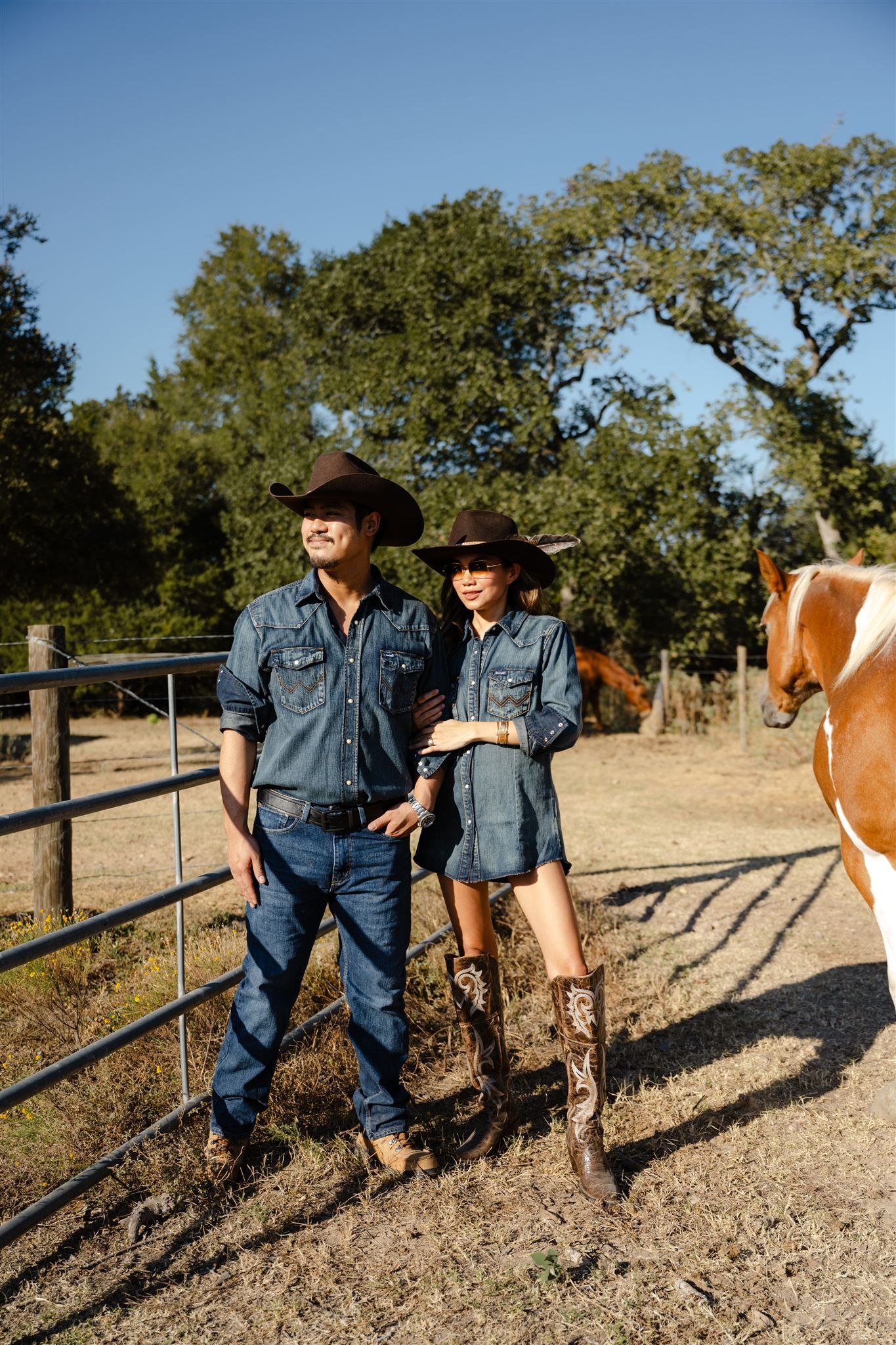 A couple in denim work clothes walking along a dirt path with cattle and oak trees in the background.
