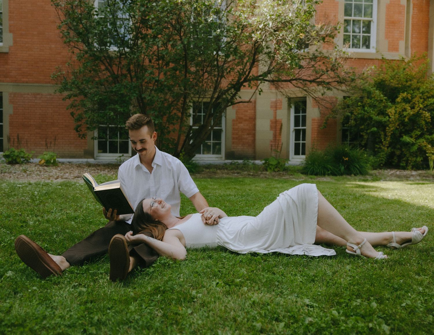 A couple lies on a grassy lawn near a brick building in a romantic outdoor photoshoot.
