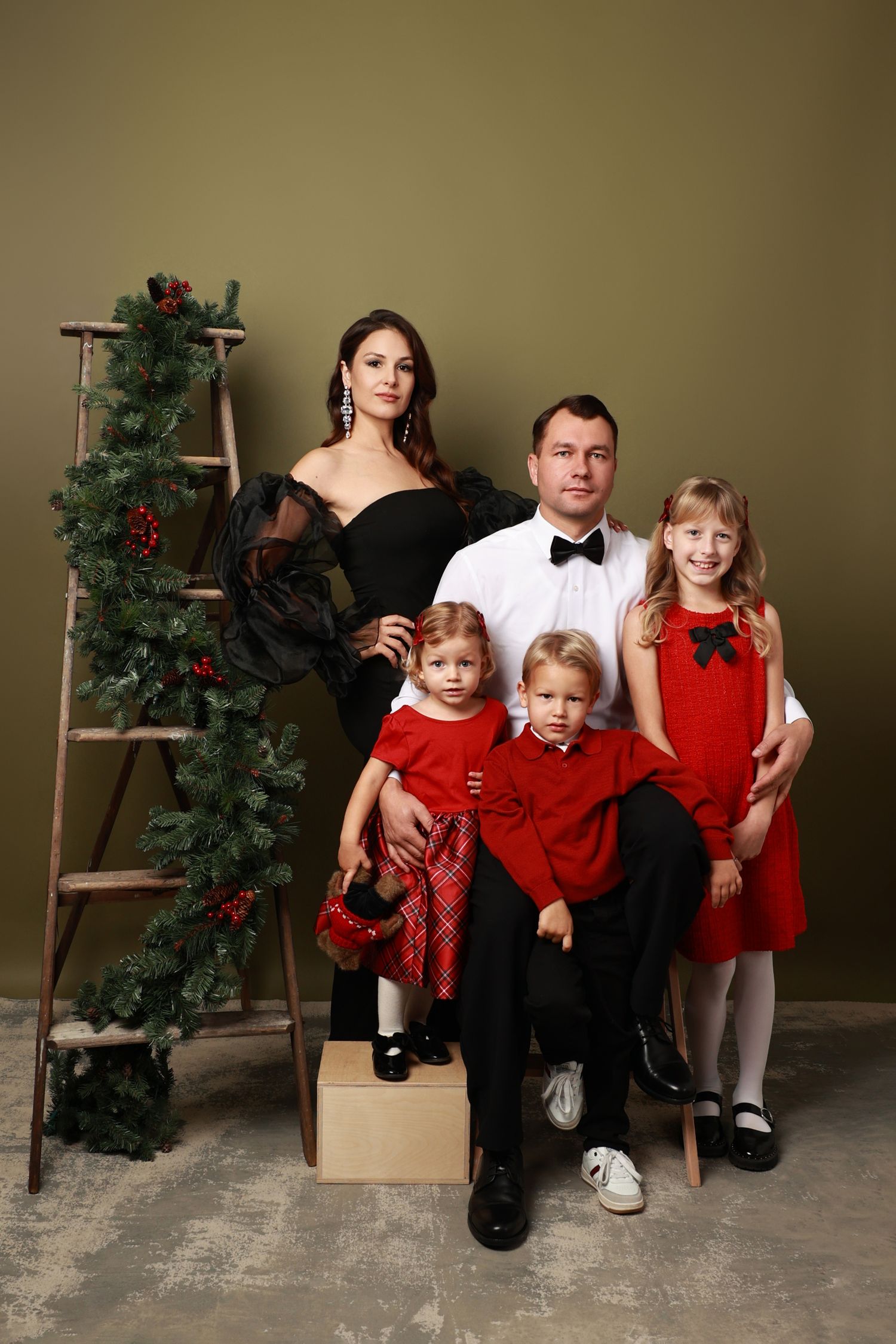 Elegant family portrait in formal attire with black and red holiday theme next to decorated ladder with garland.