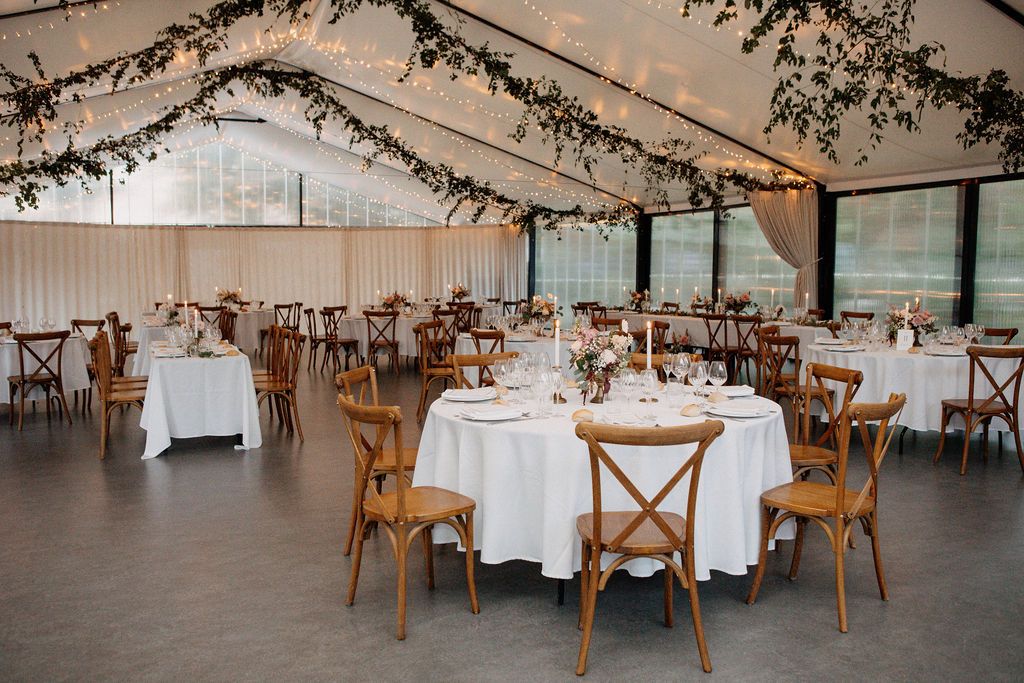 Elegant wedding reception hall with round tables, wooden cross-back chairs and fairy light ceiling decor.