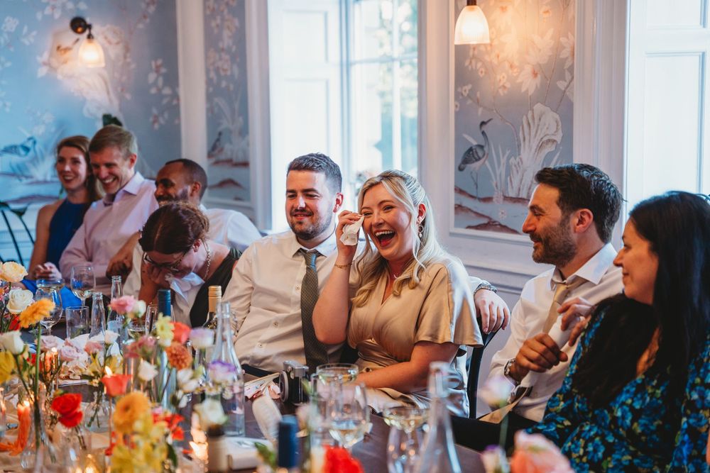 Wedding reception guests laughing and enjoying speeches during an indoor celebration.
