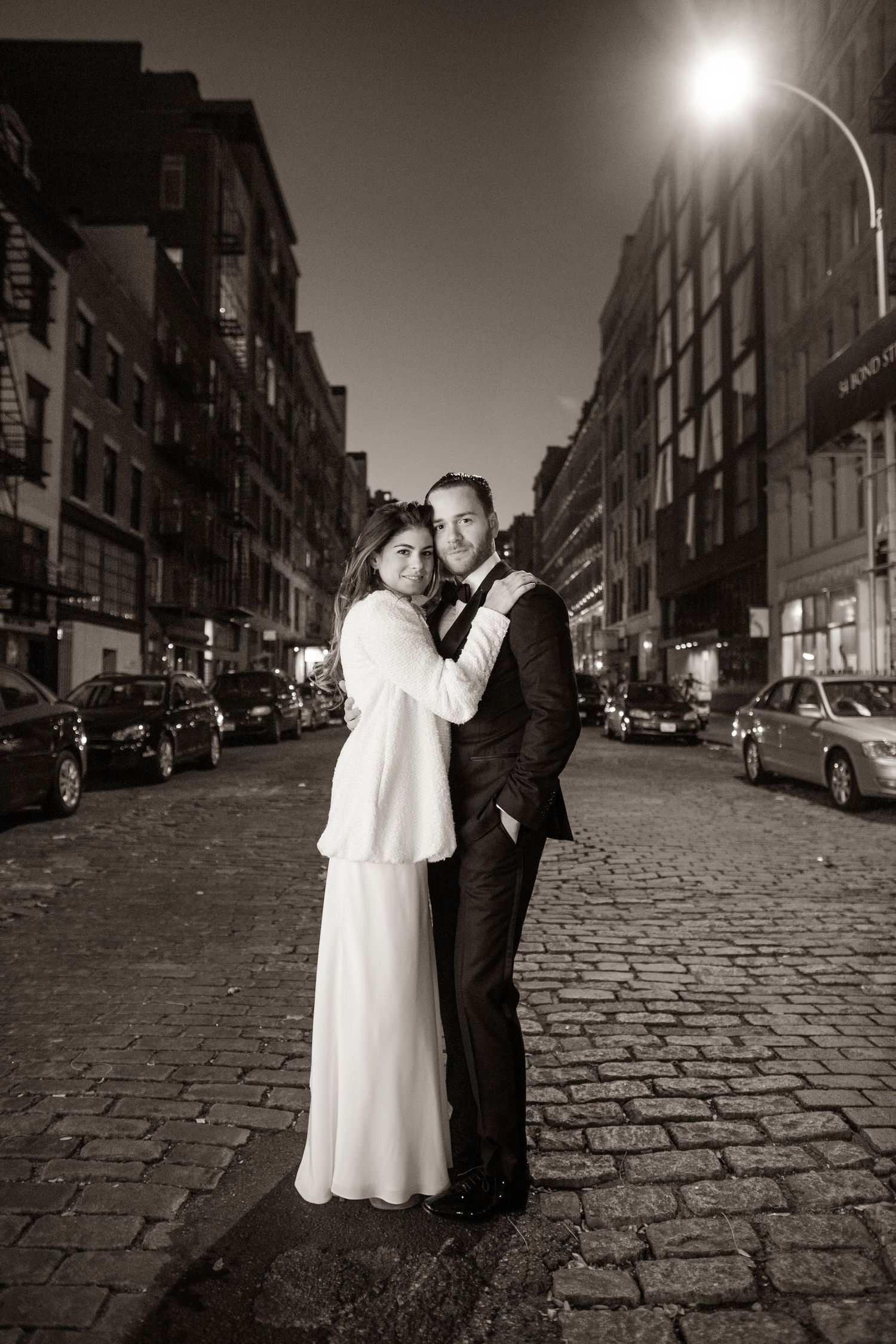 A romantic black and white photo series of a couple on a cobblestone street at night with street lamps glowing.