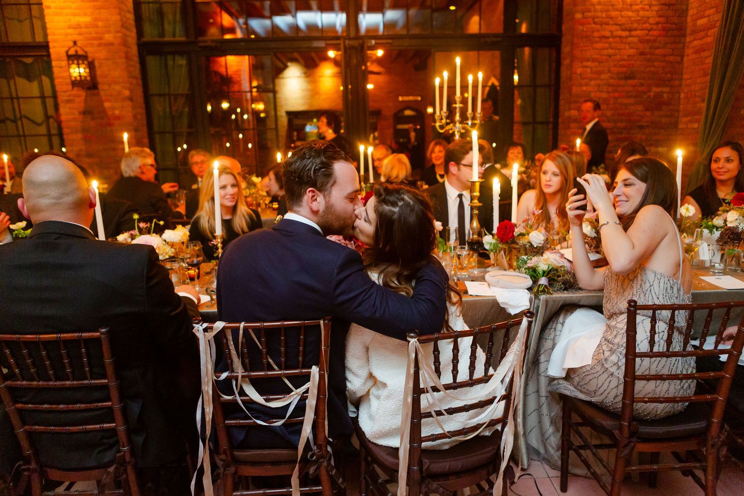 Wedding reception guests seated at long tables in a rustic brick-walled venue with warm lighting and candle centerpieces.