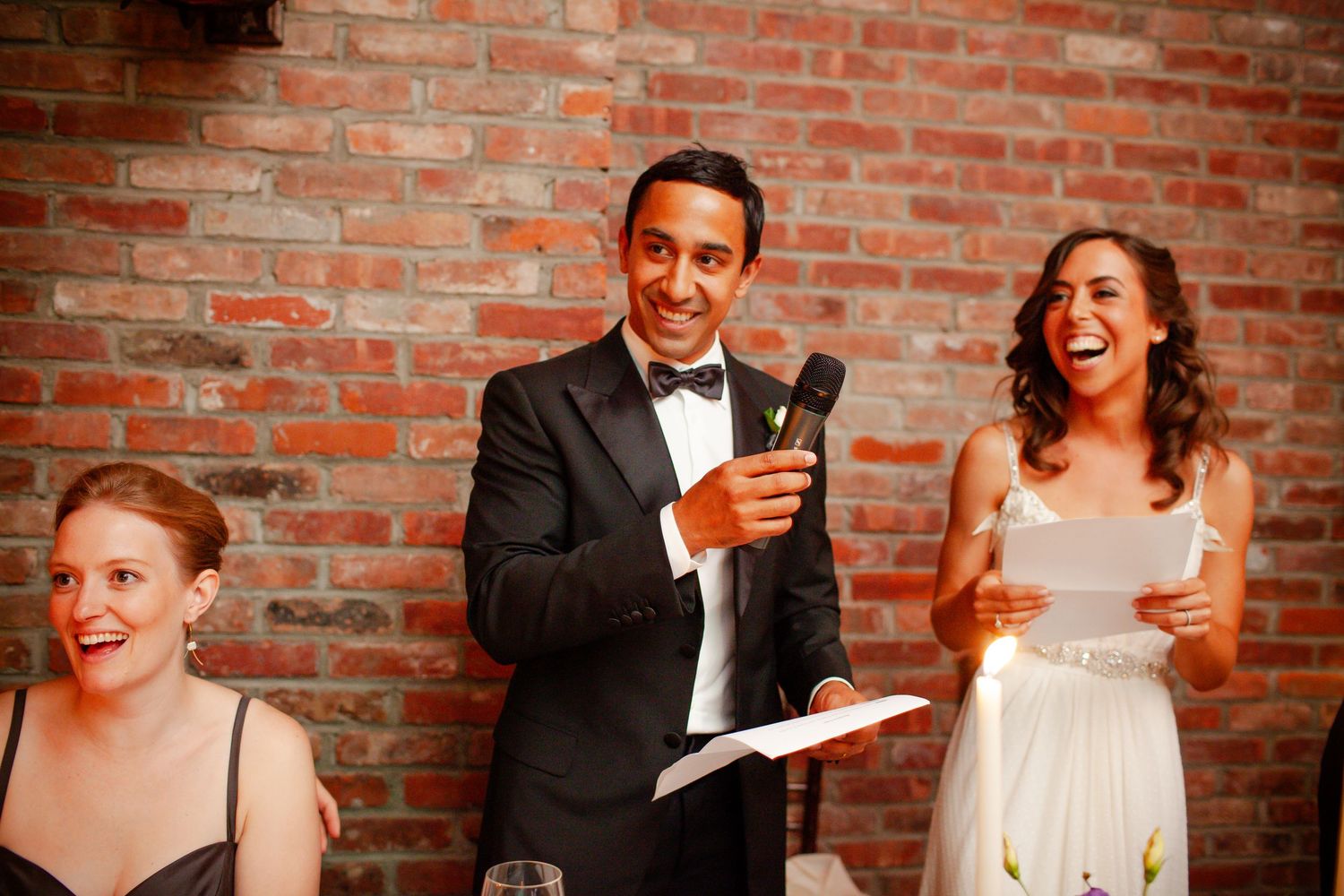 Wedding guests sharing laughter during reception speeches against exposed brick wall background.