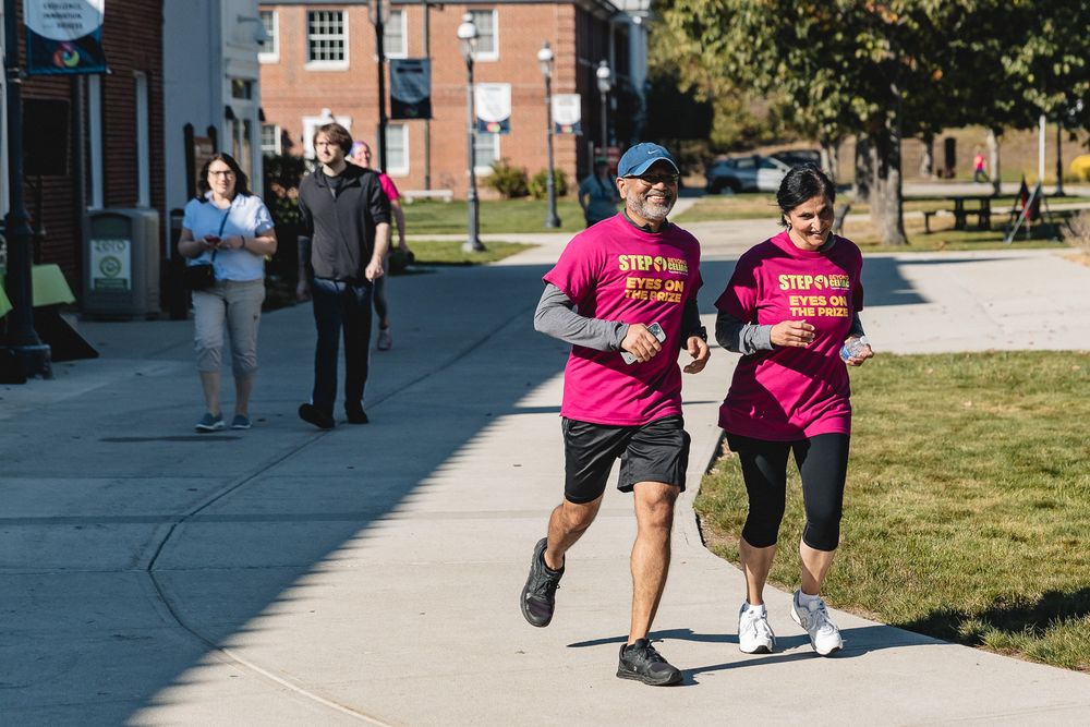 Participants in bright pink shirts run along a sidewalk during a community fitness event on a sunny day.