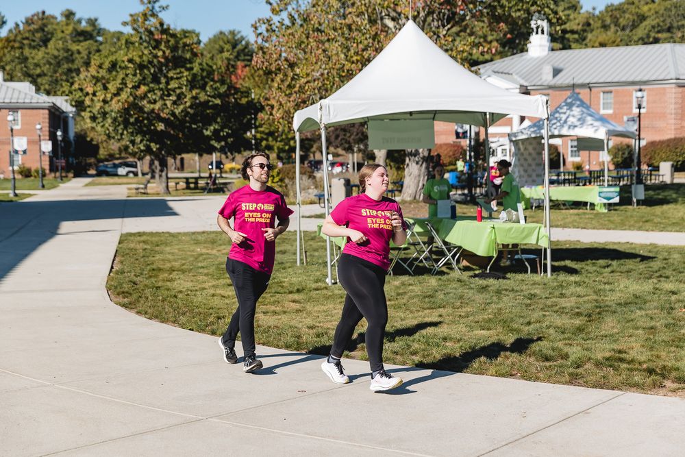 Participants in bright pink shirts running along a paved path at an outdoor community event with white tents.