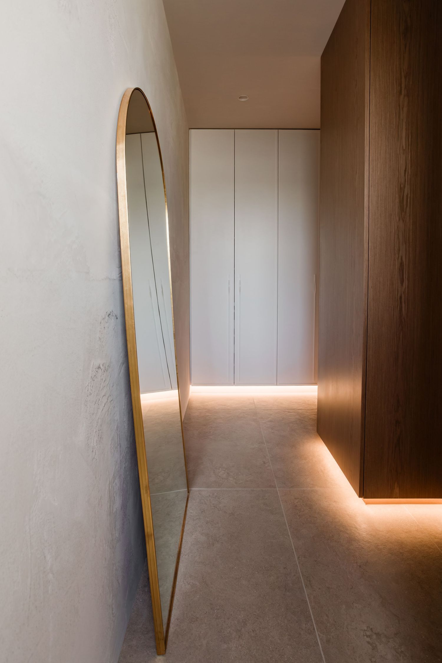 Modern hallway with oval mirror and minimalist lighting features warm ambient glow along concrete floor.