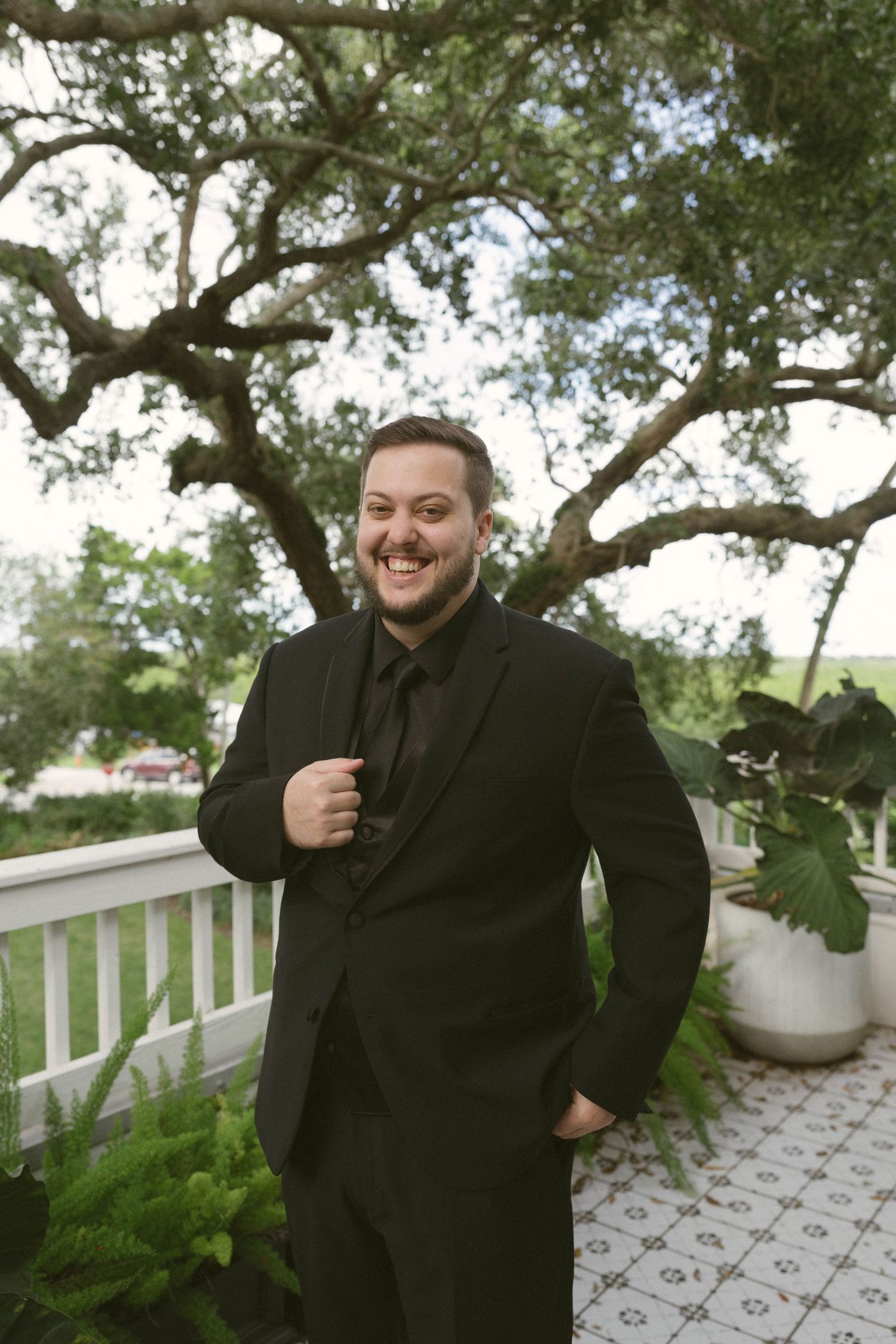 A person in black clothing sits and smiles on an outdoor porch with white railings and green plants.