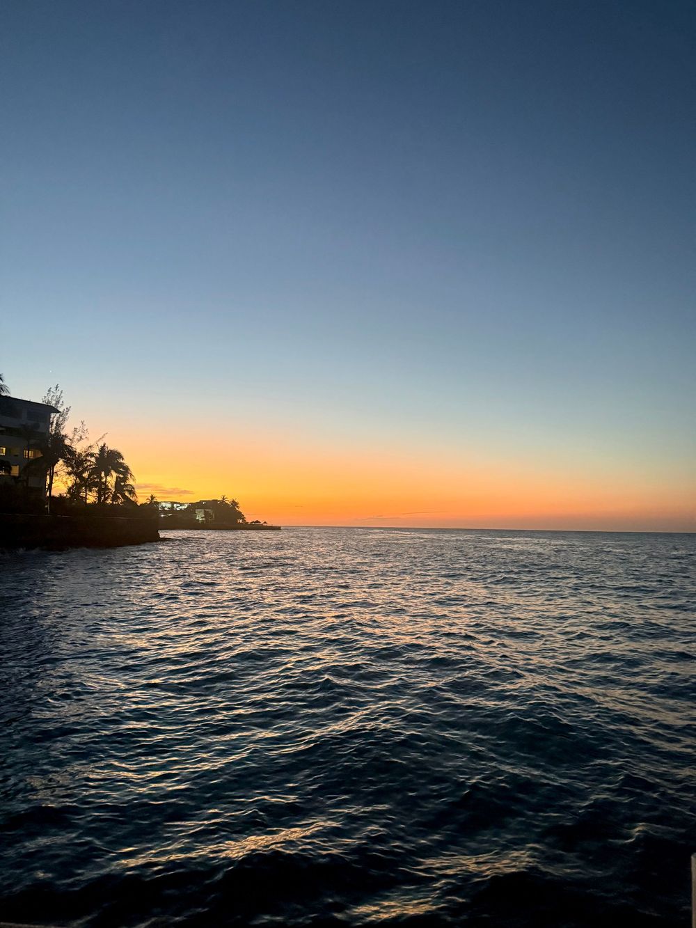 Stunning ocean sunset with vibrant orange horizon, calm waters, and silhouetted tropical buildings and palm trees.