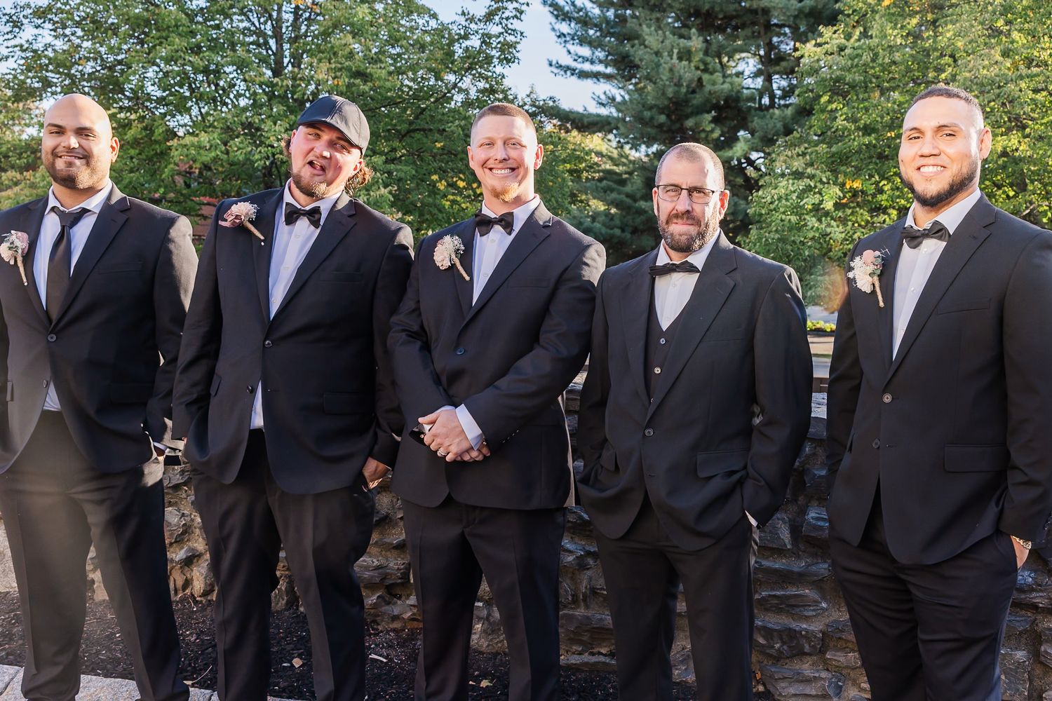A group of groomsmen in black suits and bow ties stand together outdoors for a formal wedding photo.