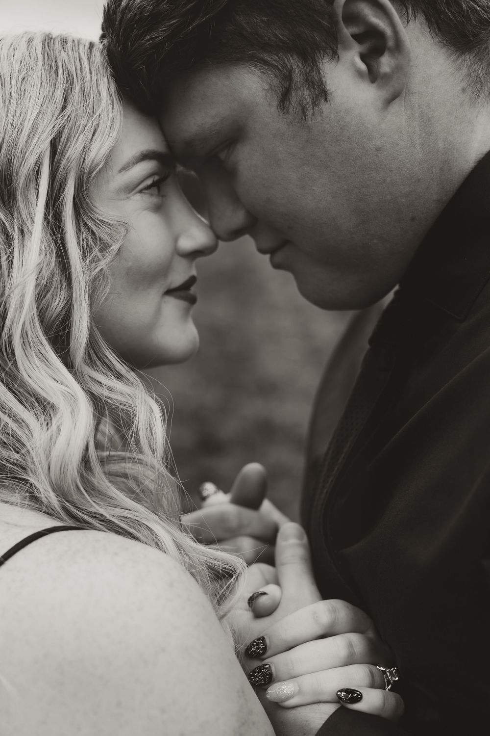Black and white close-up of a romantic couple touching foreheads, holding hands tenderly outdoors.