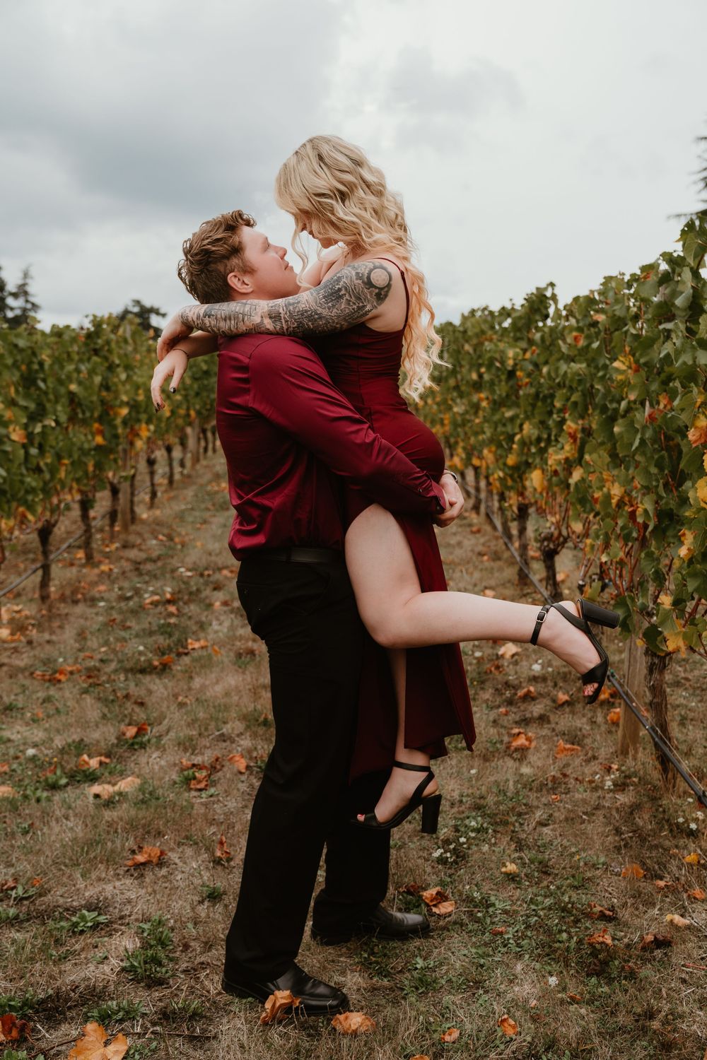 Couple in matching burgundy outfits embracing in a vineyard, man lifting woman among autumn grapevines.