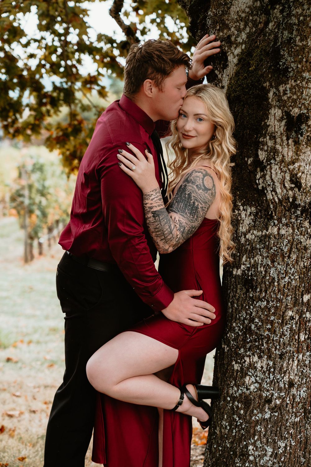 Couple embracing by a tree in vineyard, woman in red dress with tattoos, man in burgundy shirt kissing her.