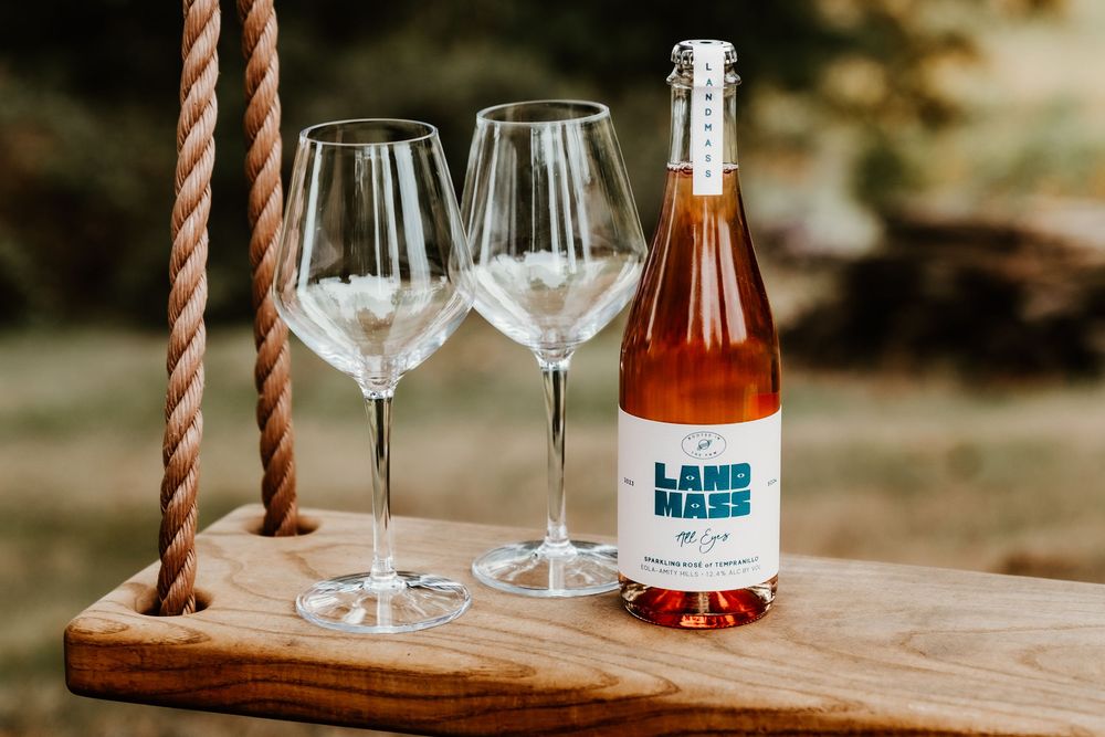 Bottle of Landmass rose wine with two empty wine glasses on a wooden swing outdoors.