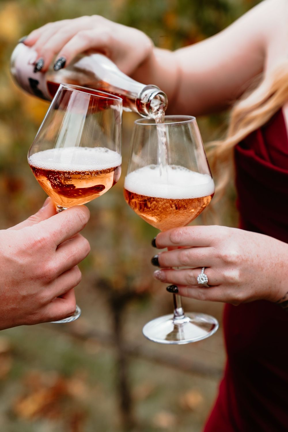 Close-up of rose wine being poured into glasses held by couple with engagement ring visible.