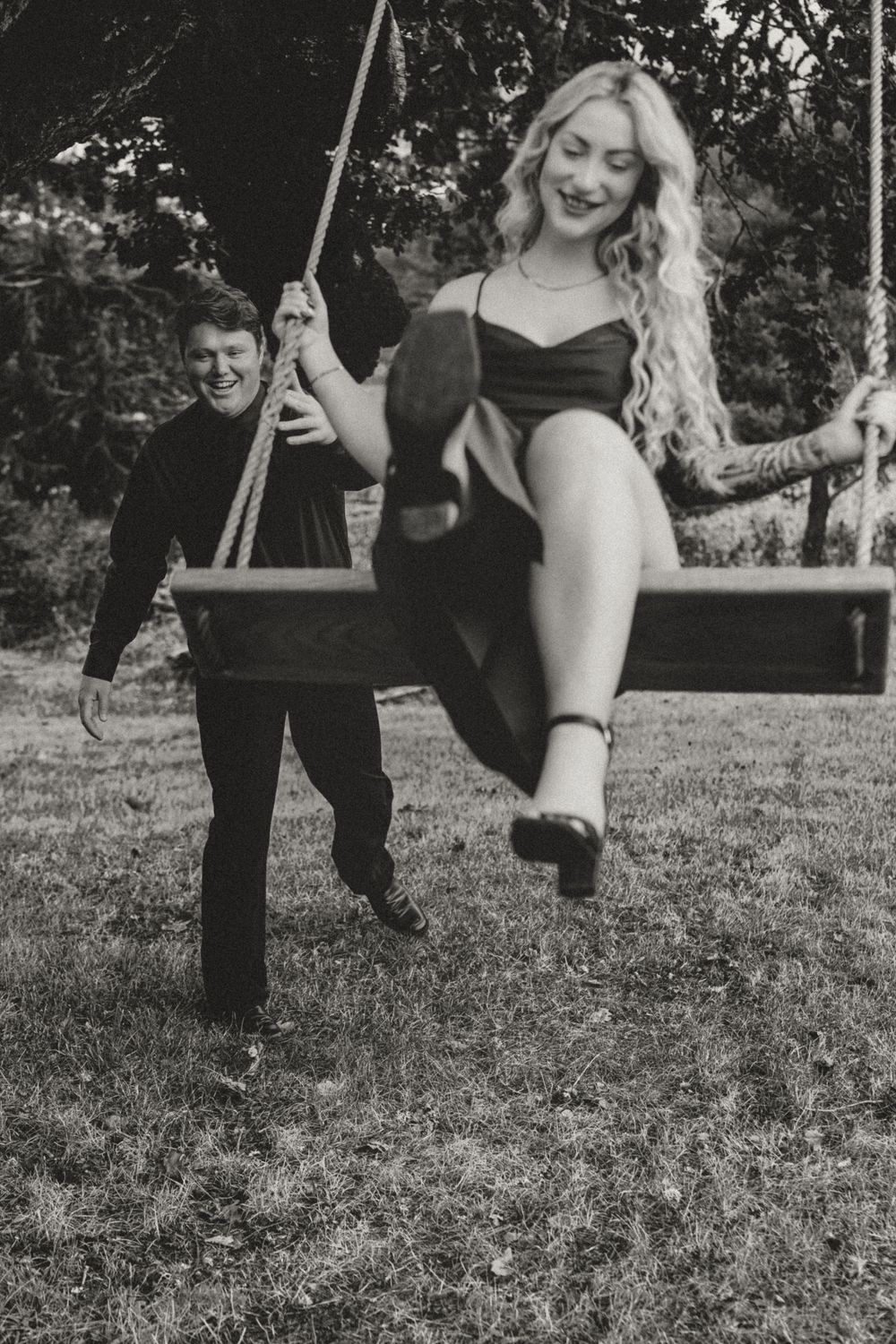 Black and white photo of a laughing woman on a swing being pushed by a smiling man outdoors.