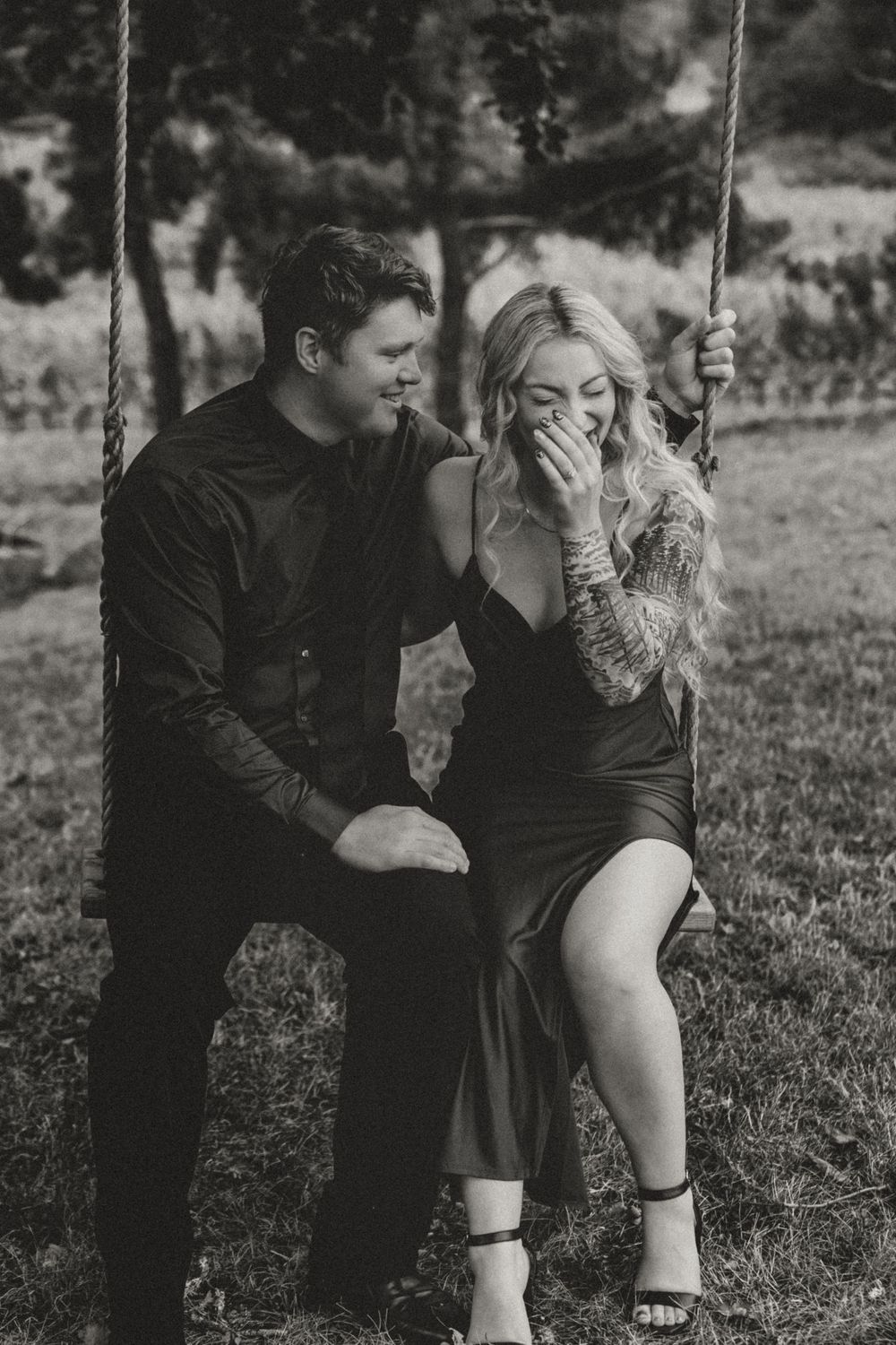 Black and white photo of a couple sitting on a swing, woman laughing while man whispers to her.