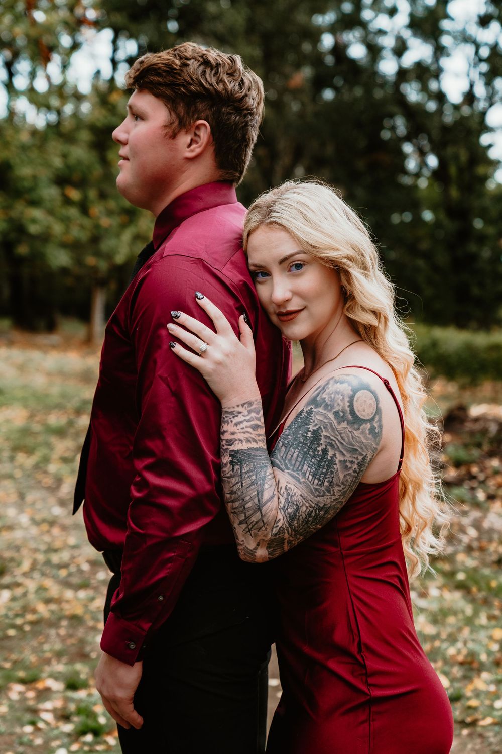 Woman with sleeve tattoo in red dress embracing man in burgundy shirt from behind in autumn forest.