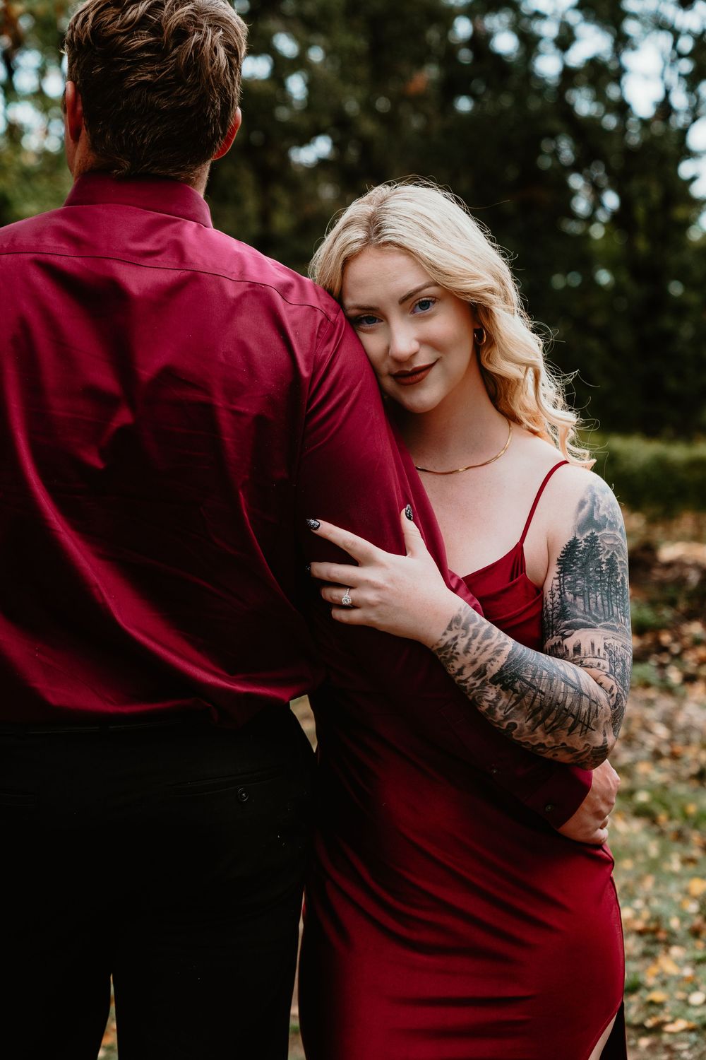 Tattooed woman in red dress embracing man in burgundy shirt from behind in outdoor autumn setting.