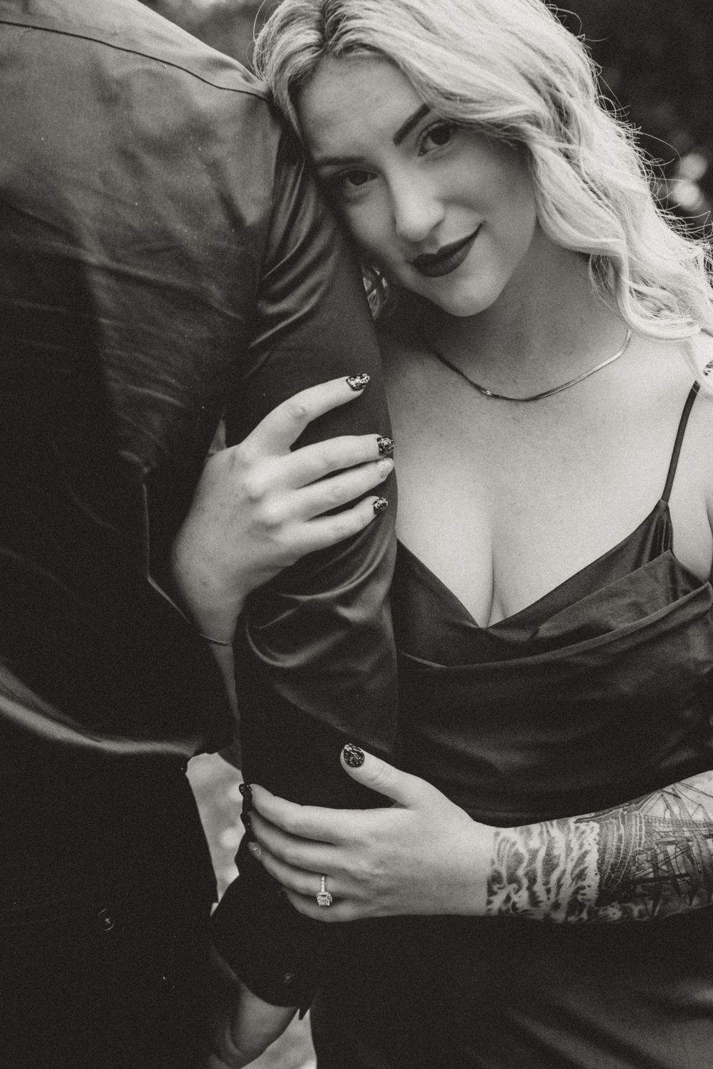 Black and white intimate portrait of tattooed woman in satin dress embracing her partner outdoors.