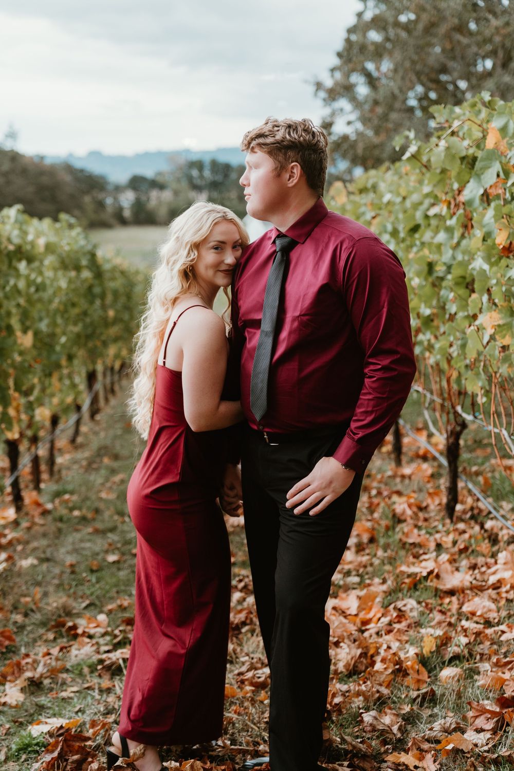 Couple posing in autumn vineyard, woman in red dress smiling, man in burgundy shirt and black tie.