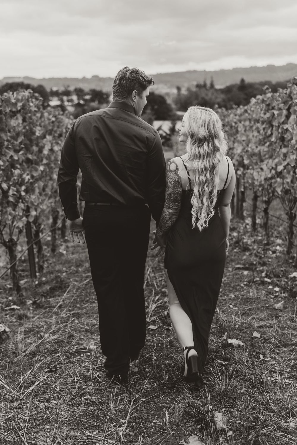 Black and white photo of a couple walking hand in hand through a vineyard, woman in black dress with tattoos.