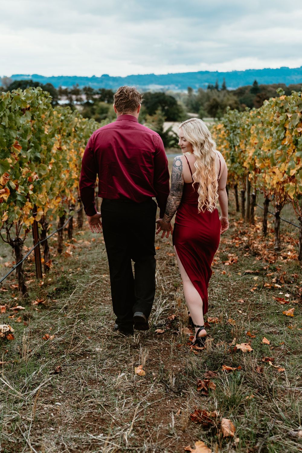 Couple walking hand in hand through colorful autumn vineyard rows, woman in red dress with back tattoo.