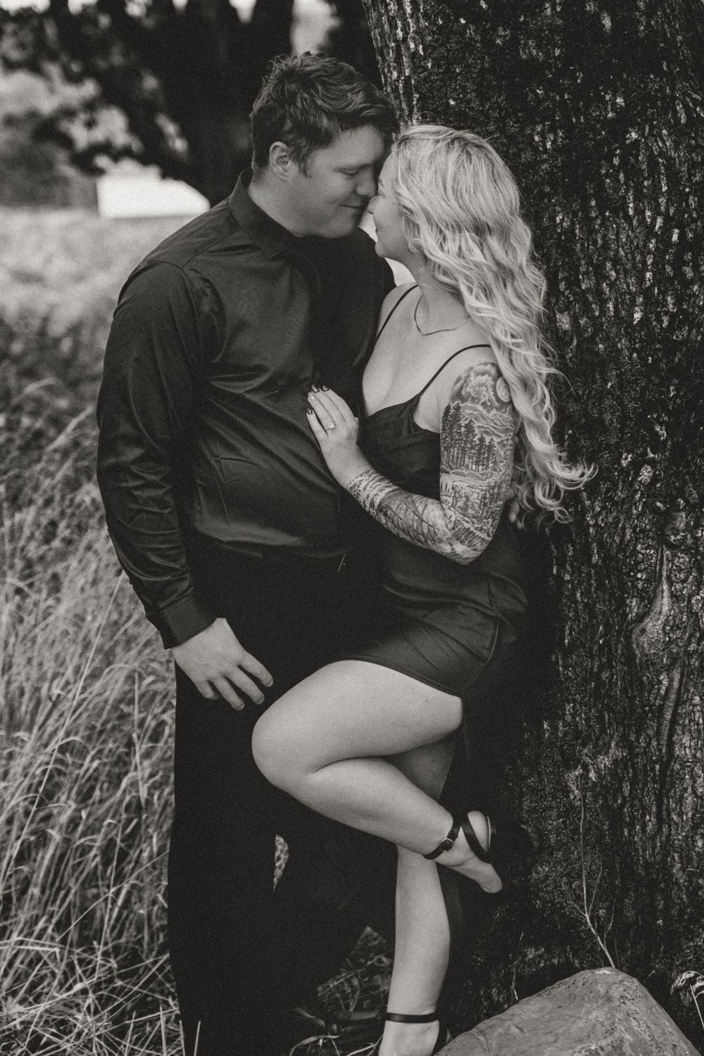Black and white photo of a couple sharing a romantic moment against a tree, woman with blonde hair and sleeve tattoos.