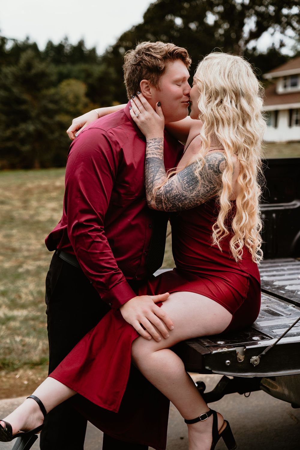 Couple sharing a kiss outdoors, woman with tattoos in red dress sitting on truck tailgate, man in crimson shirt.