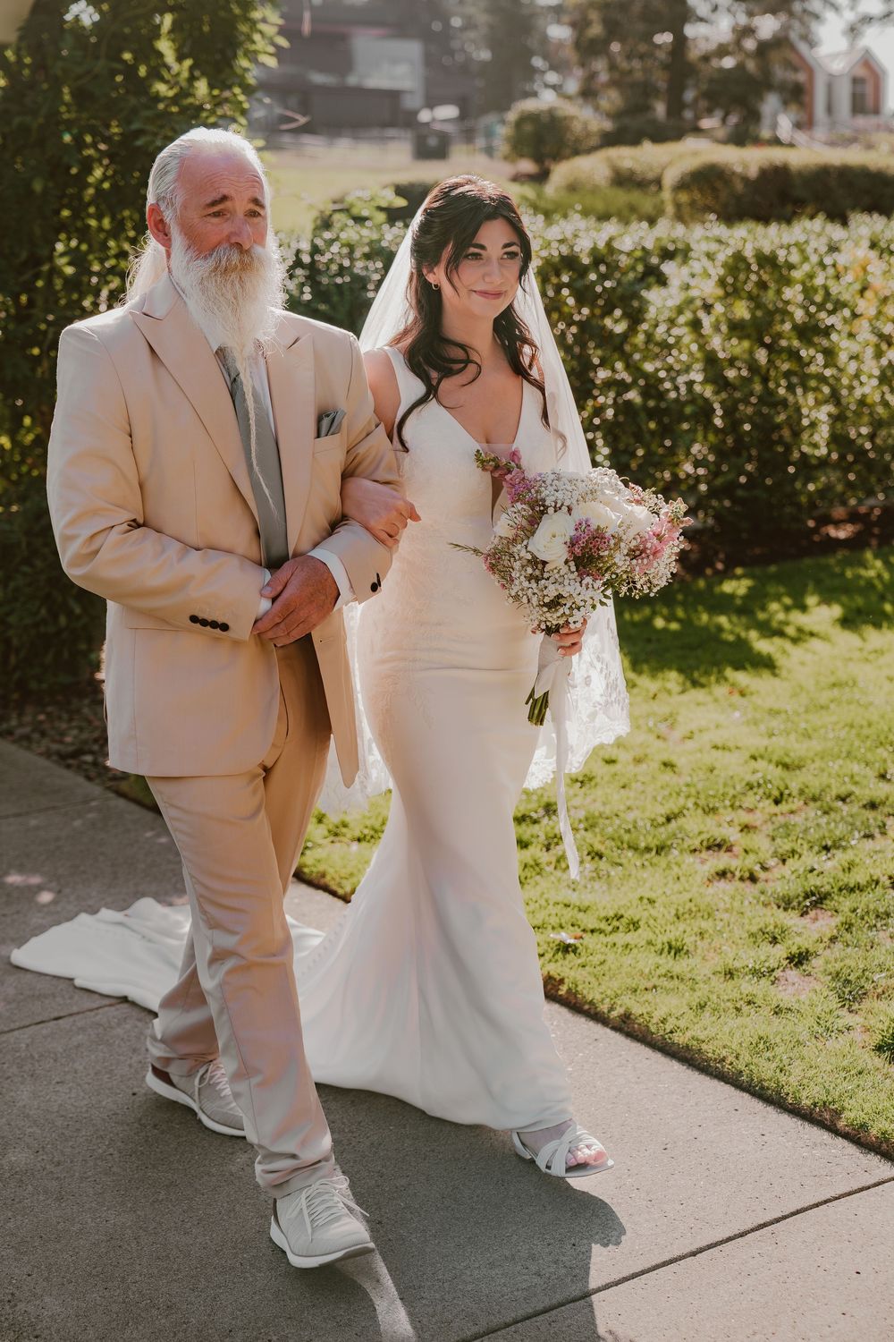 Bride in fitted white gown holding bouquet walks with bearded father in beige suit.