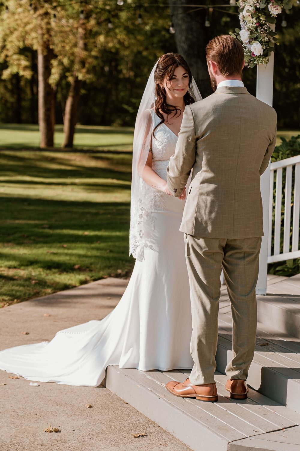 Smiling bride in lace veil holds hands with groom during outdoor wedding ceremony on sunny day.