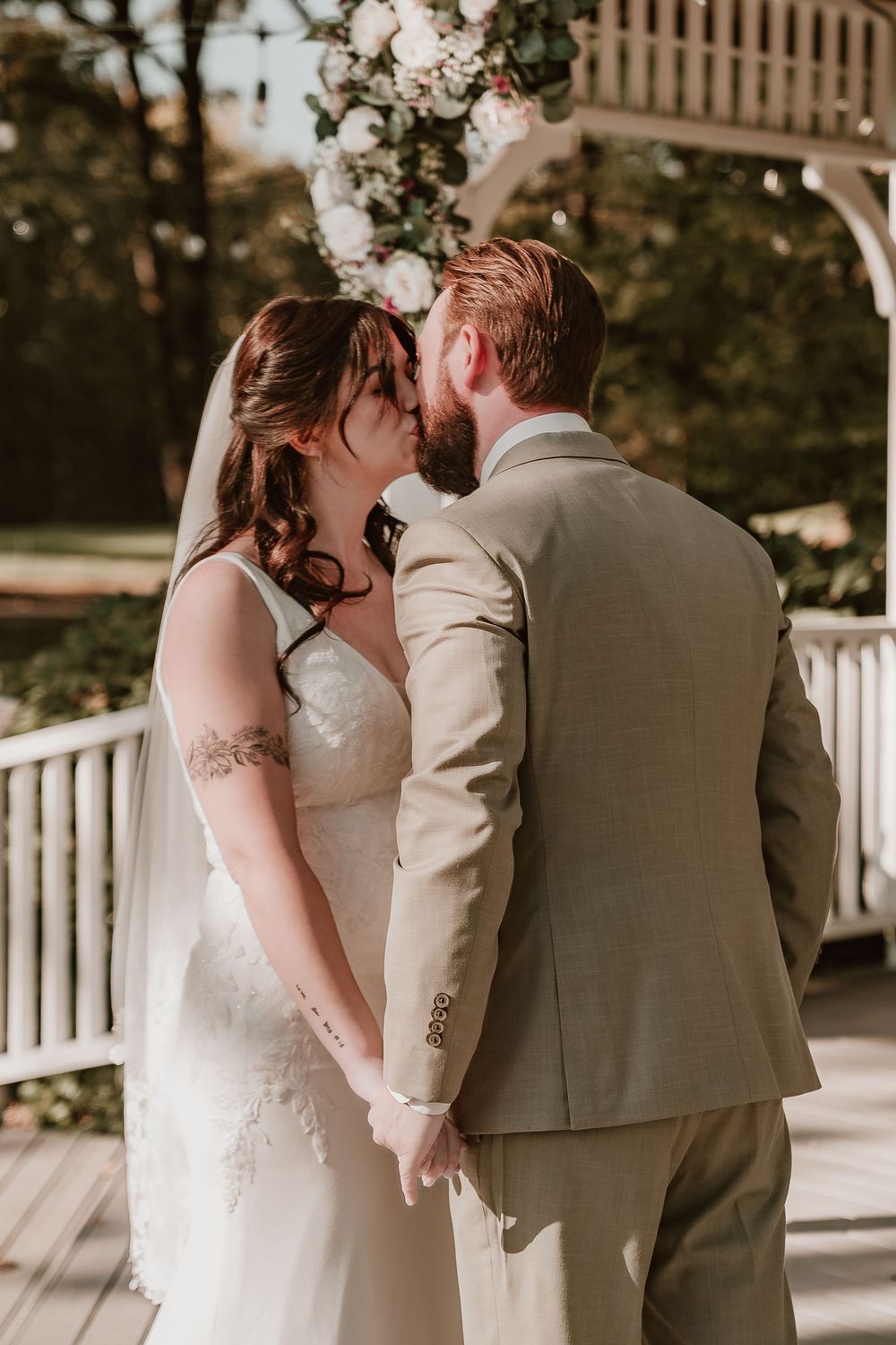 Bride and groom share a tender kiss at outdoor wedding ceremony near white gazebo with floral arch.