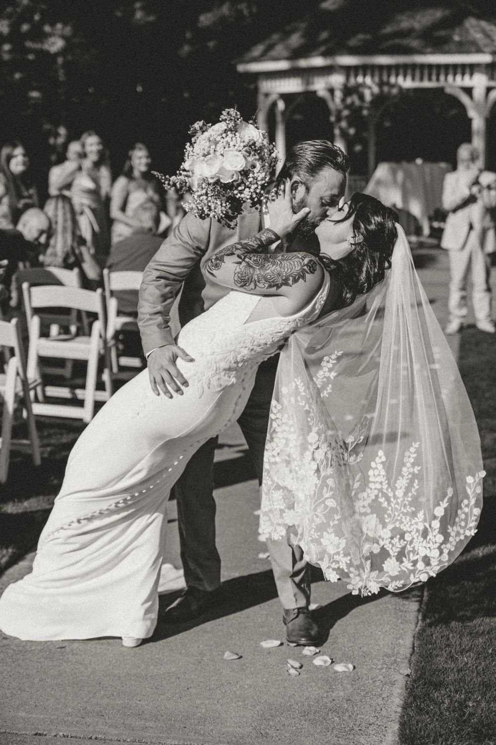 Groom dips bride for romantic kiss while walking down aisle at outdoor wedding ceremony, black and white photo.