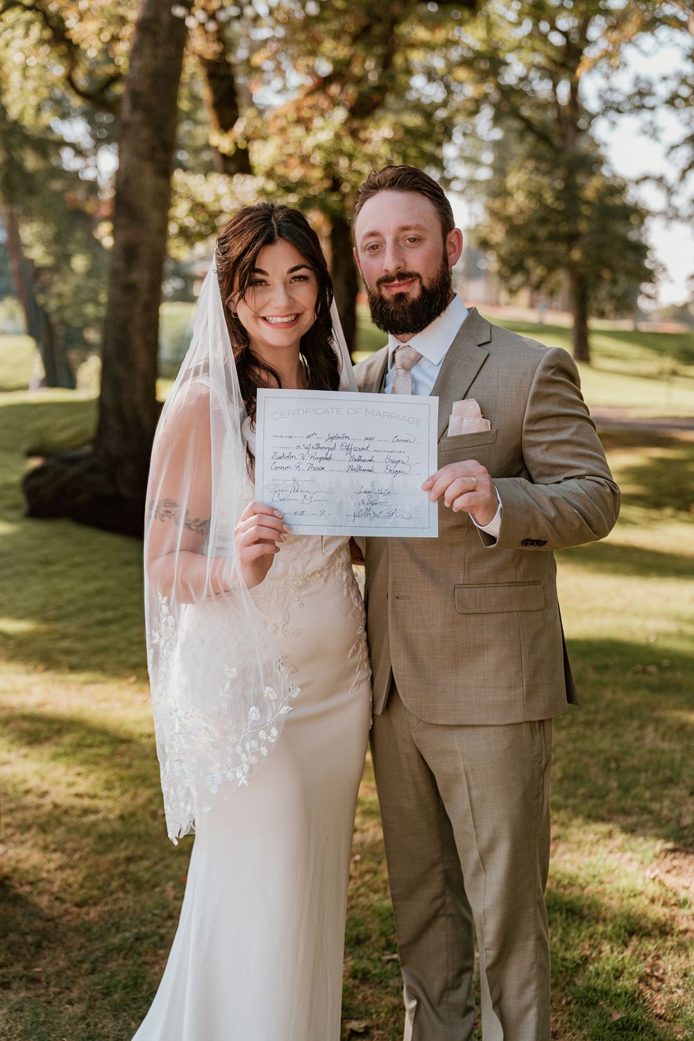 Happy newlyweds smile while holding their signed marriage certificate outdoors on their wedding day.