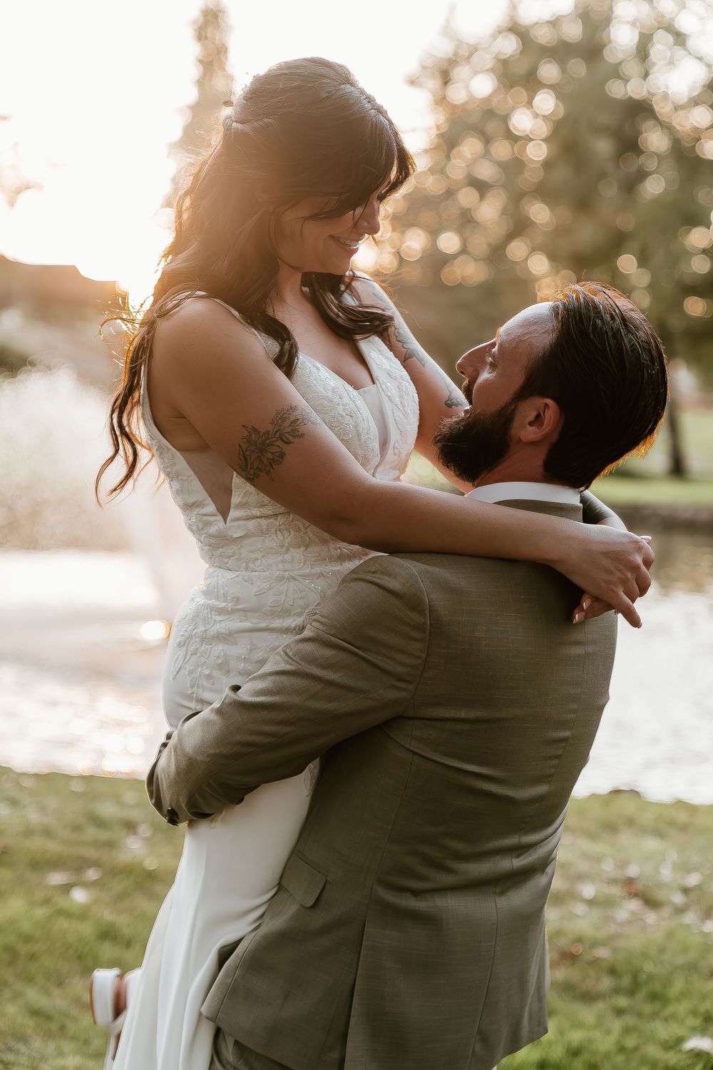 Groom lifts bride in white lace gown during golden hour wedding portraits near scenic pond.