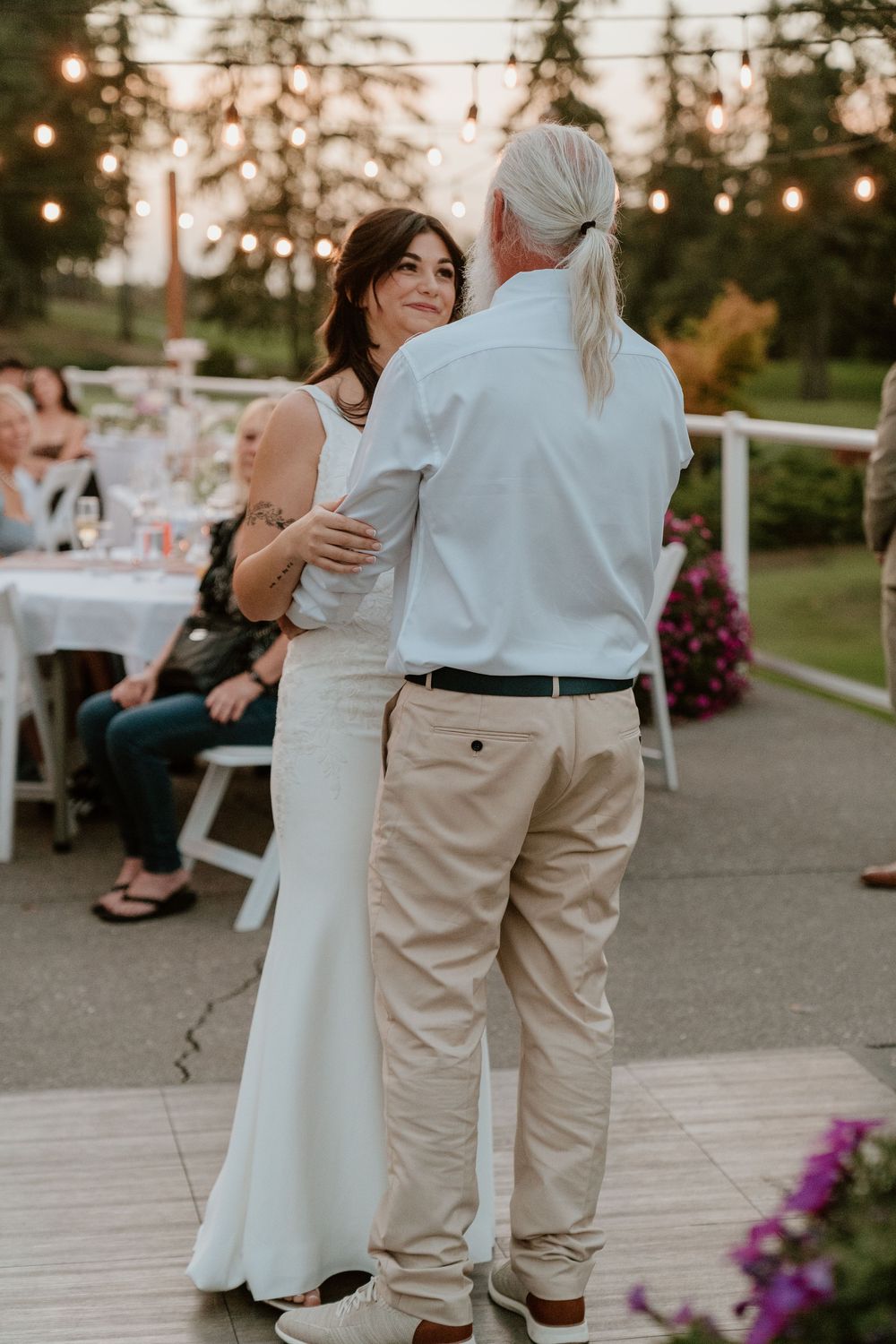 Bride in white gown shares father-daughter dance at outdoor wedding reception with string lights.