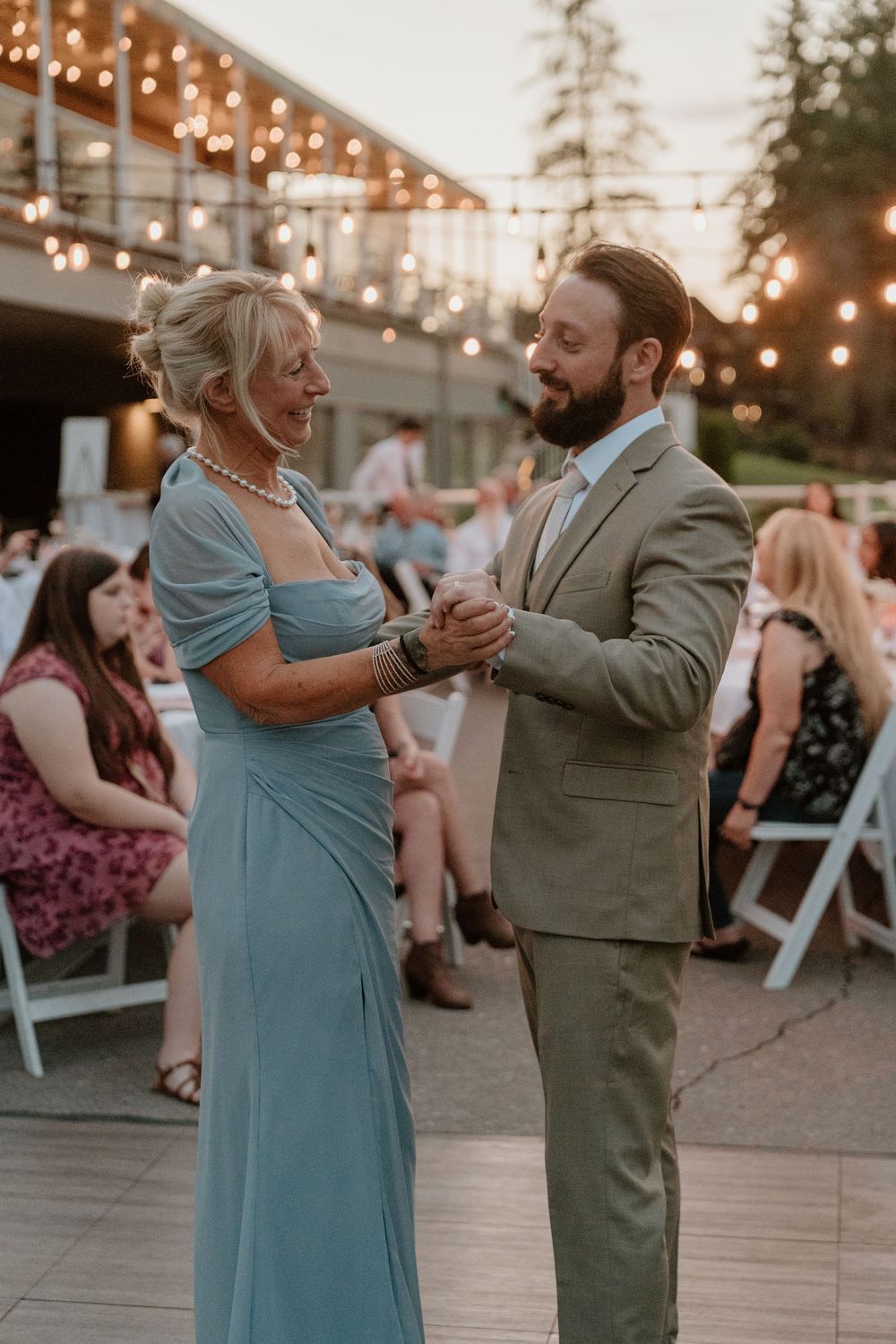 Groom in tan suit shares mother-son dance with woman in blue gown at outdoor wedding reception with string lights.