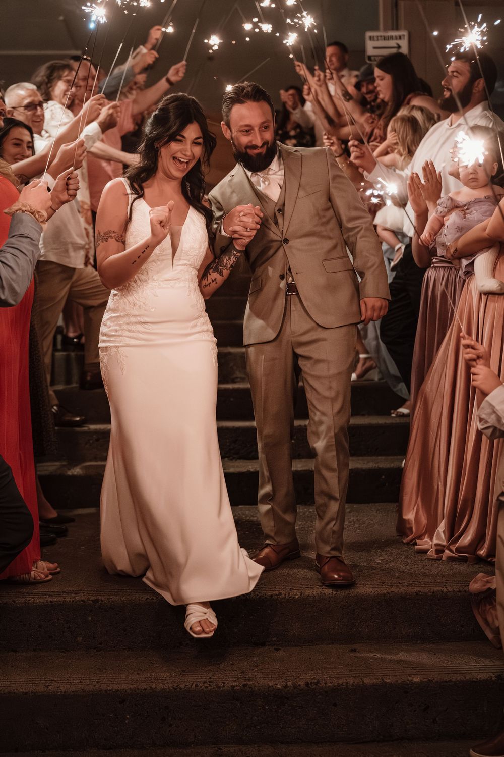 Newlywed couple exits wedding ceremony through sparkler tunnel, bride in white gown and groom in tan suit.