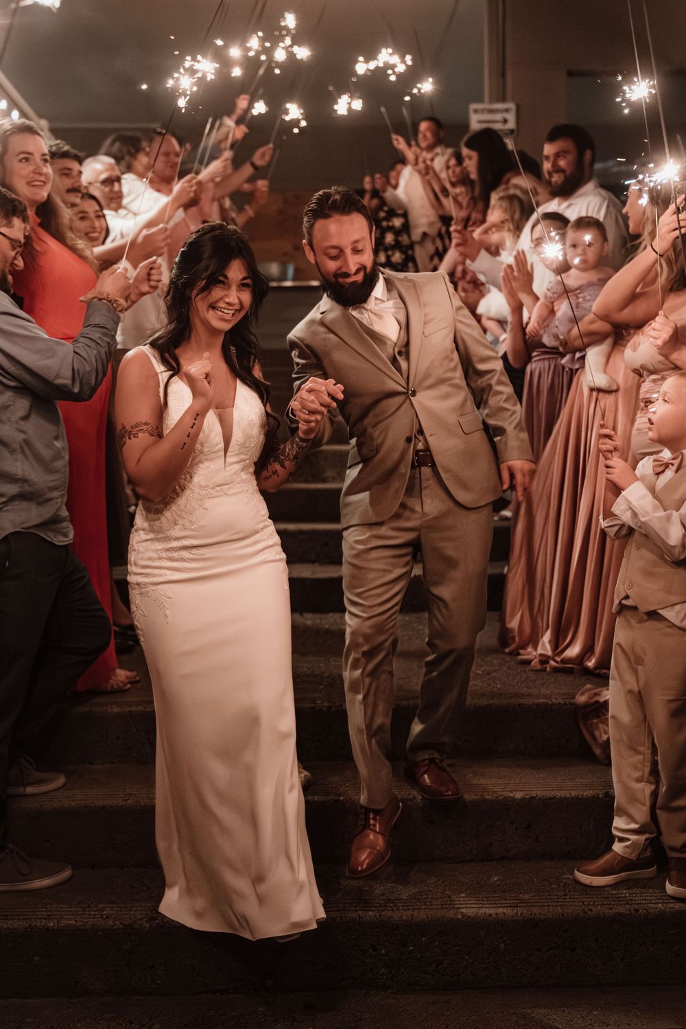 Smiling bride and bearded groom in tan suit walk through sparkler send-off surrounded by cheering wedding guests.
