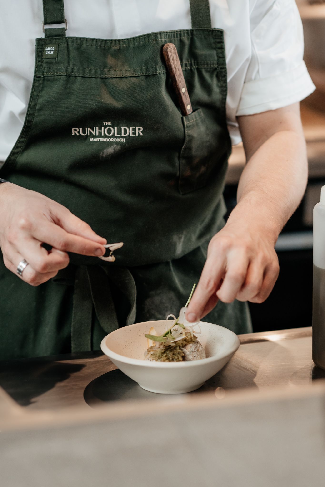Chef wearing green Runholder apron garnishes a dish in white bowl while working in restaurant kitchen by August and Vine
