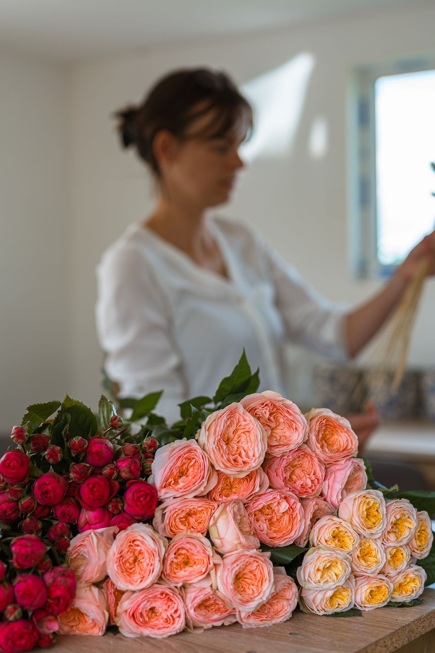 Fresh peach garden roses and vibrant red flowers arranged in a line on a wooden table with someone working in background.