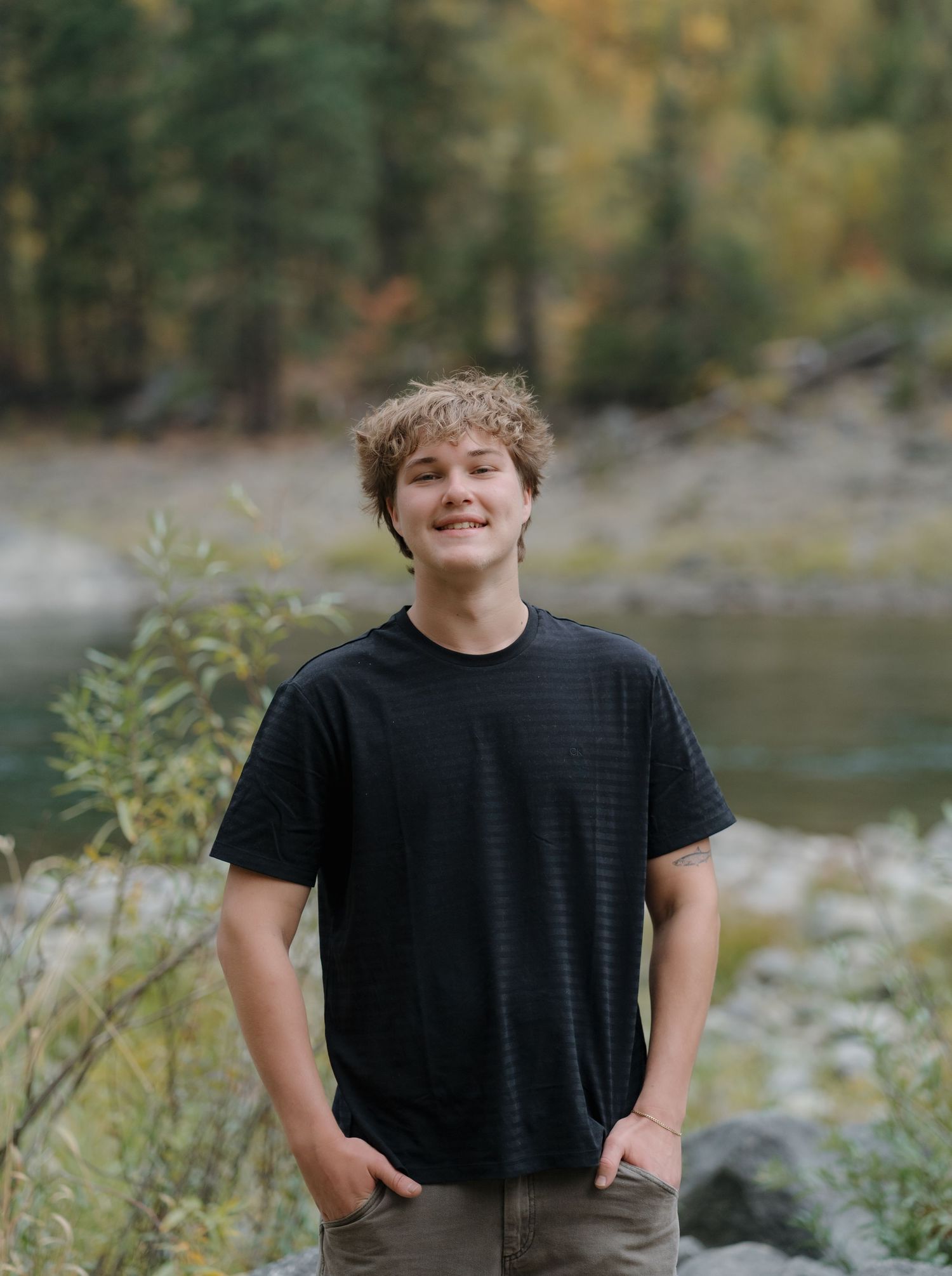 Person in black t-shirt sits outdoors near a lake surrounded by autumn foliage and natural scenery.