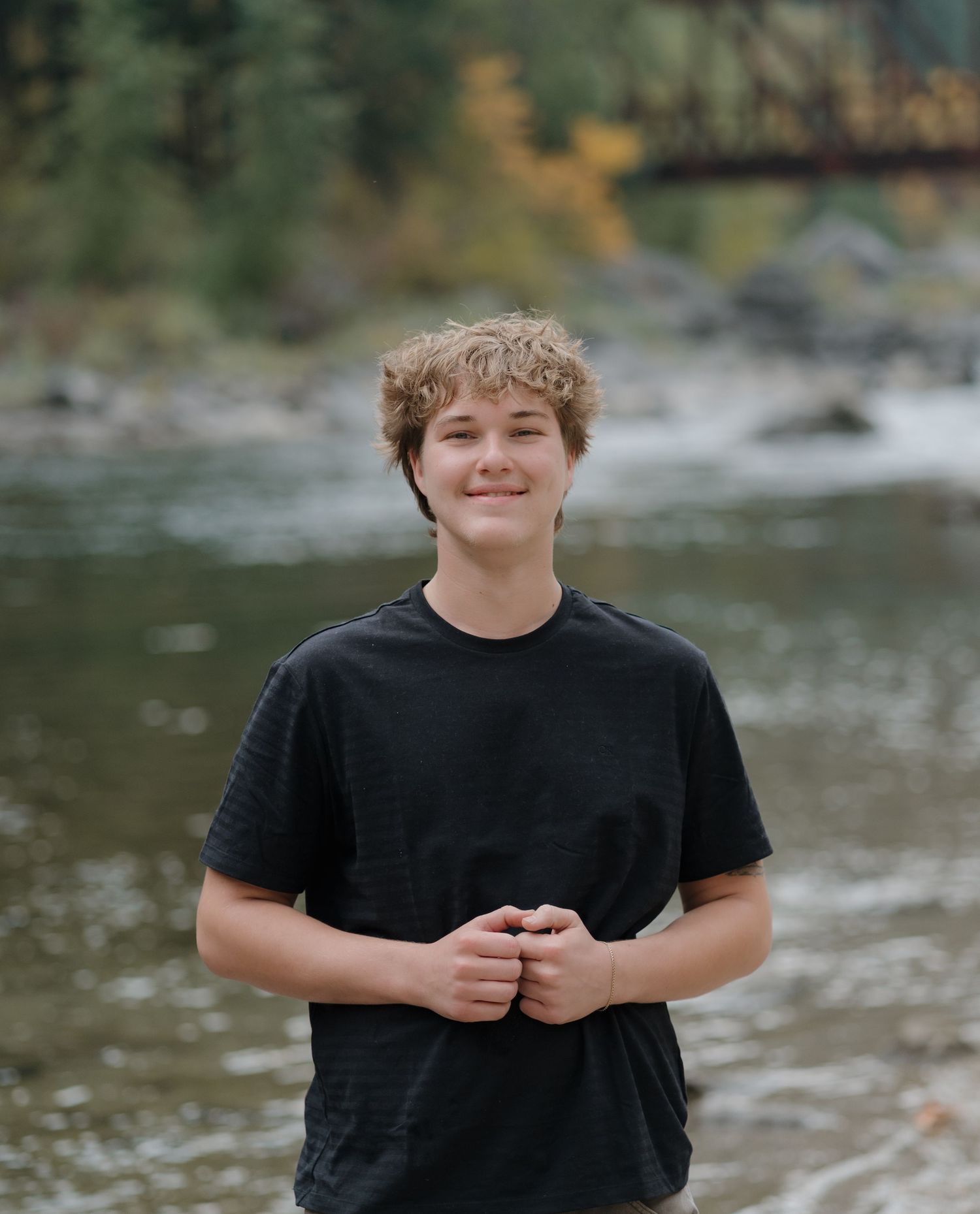 A person in a black t-shirt stands near a rocky riverbank surrounded by forest.