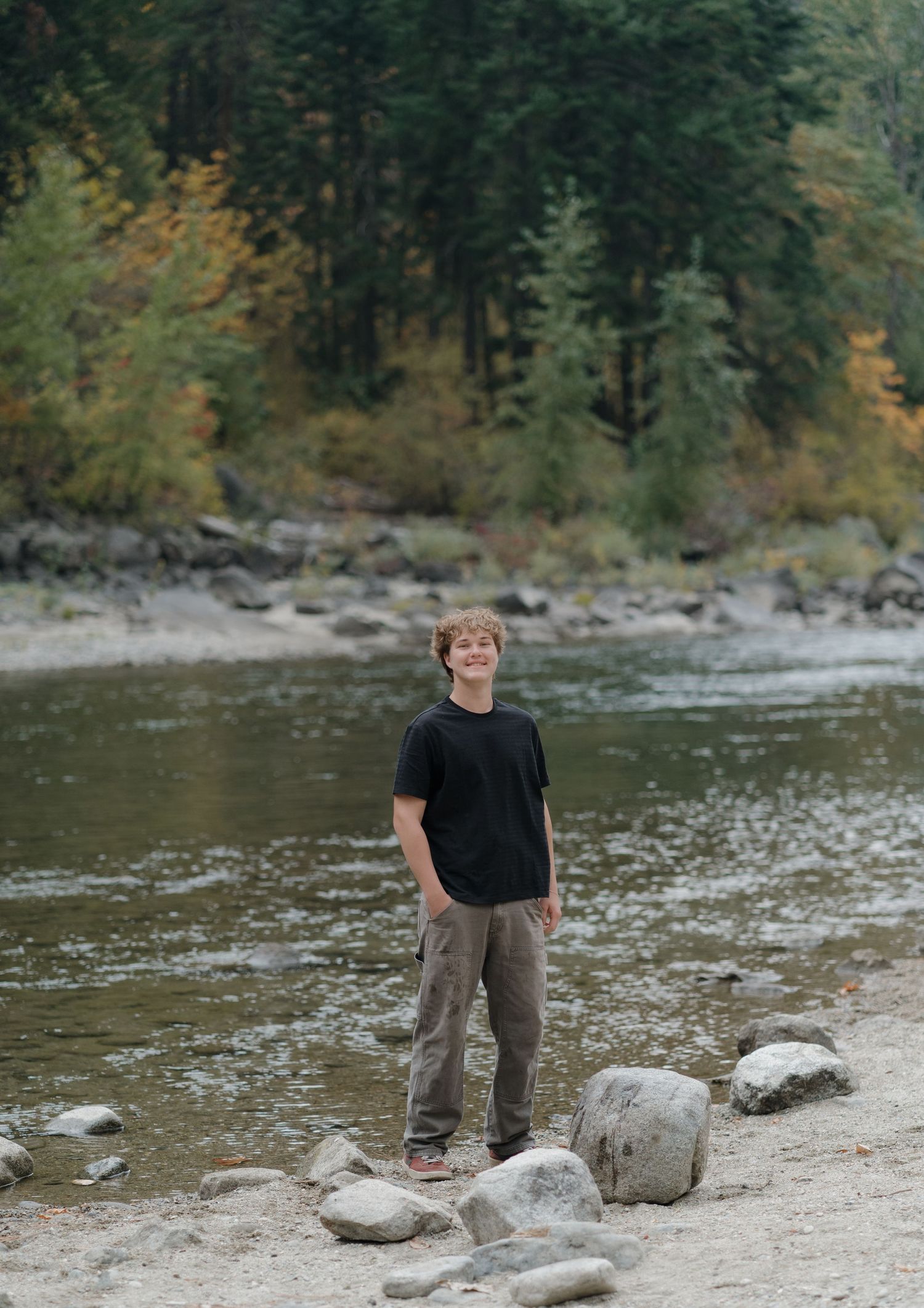 A scenic riverbank with rocks and trees where someone stands against a forested background.