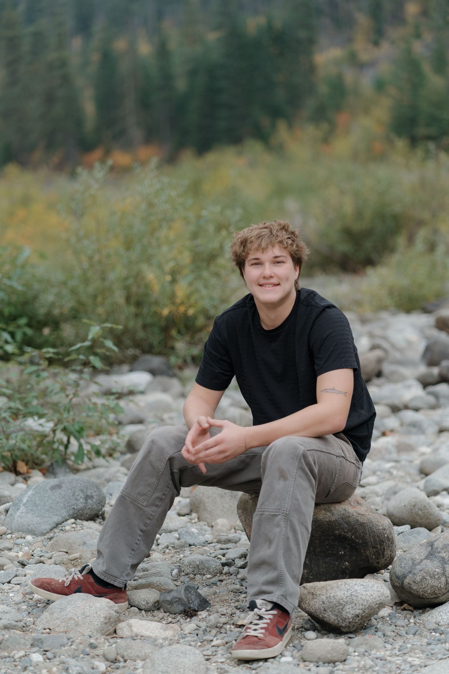 A person in a black shirt and gray pants sits casually on rocky ground surrounded by natural scenery.