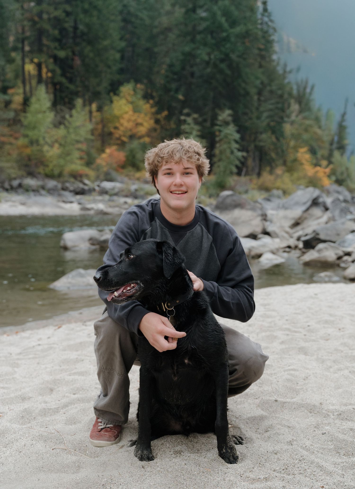 A black labrador retriever sits with its companion on a rocky riverbank surrounded by evergreen trees and autumn foliage.