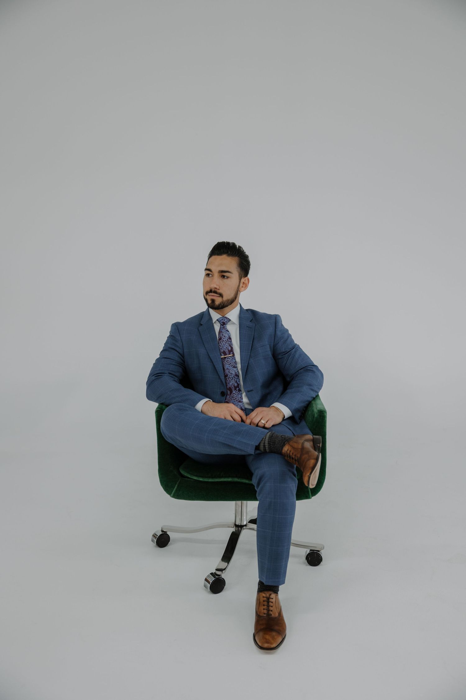 Professional wearing a navy blue suit and floral tie sits confidently in a green office chair against white backdrop.