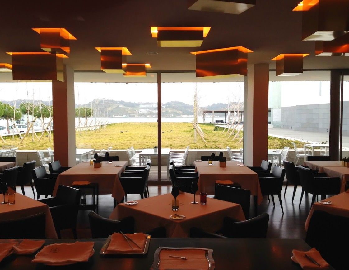 A modern restaurant dining area with orange lighting fixtures and black chairs overlooking large windows with outdoor views.